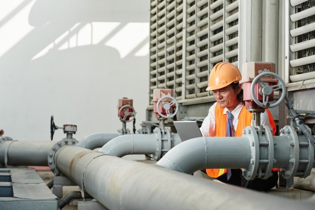 An engineer using a laptop beside large industrial pipes and control valves.