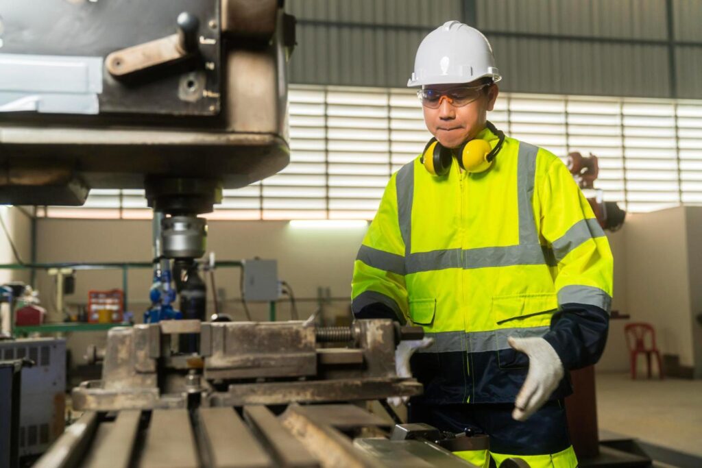Operator in a hi-vis jacket and hard hat adjusting a vertical drilling machine in a gearbox machining workshop.
