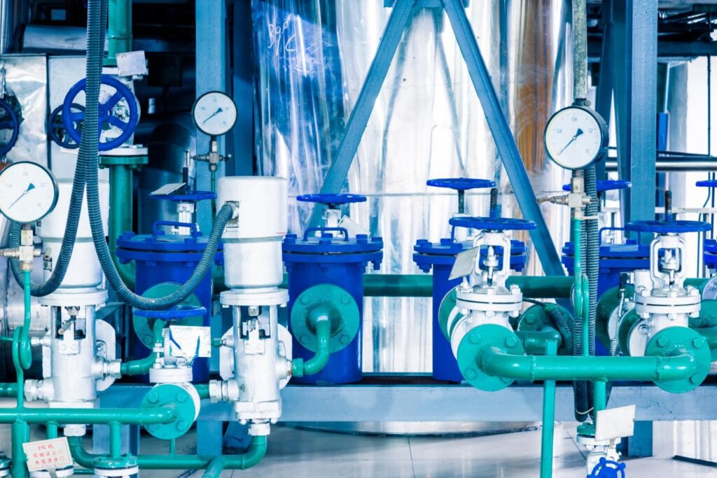 Pump room with blue centrifugal pumps, pressure gauges and handwheel valves on green and blue industrial pipework.